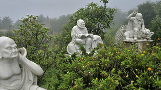 Stone statues of three figures in a lush garden, one reading, another deep in thought. Misty background with greenery and yellow flowers.