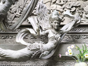 Intricate stone carving of a deity in flowing attire, adorned with ornate jewellery, set against a detailed floral background with pink and yellow flowers.