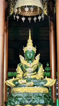 Emerald green Buddha statue adorned with intricate gold and jeweled garments, seated under an ornate golden canopy, in a dim temple.