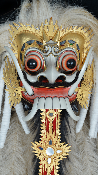 Ornate Balinese mask with big red eyes, gold accents, and white teeth on a black background. Feathers and checkered fabric surround it.