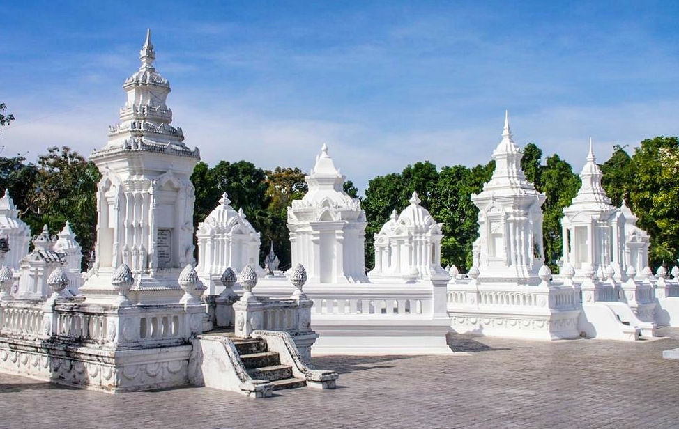 White ornate stupas under a clear blue sky, surrounded by lush green trees. The scene is peaceful and the architecture intricate.