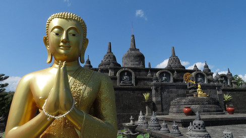 Golden Buddha statue with praying hands in front of a stone temple, under a clear blue sky. Peaceful and serene setting.