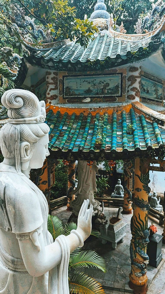 Stone statue in serene pose near ornate temple with turquoise tiles and lush greenery, evoking peace and harmony.