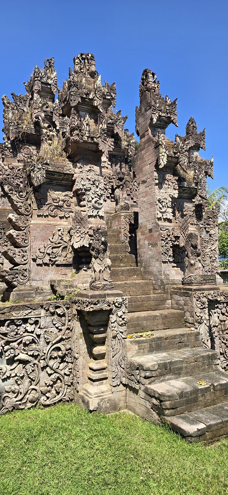 Ornate stone temple gate with stairs under a clear blue sky; small sign reads ATTENTION VISITORS, serene and sunlit.