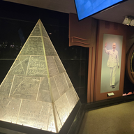 Illuminated pyramid with engraved text in various languages, next to a portrait of a smiling man in white waving, set in a dimly lit room.