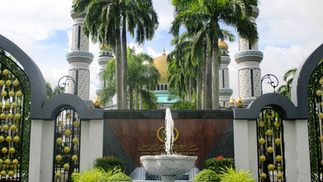 Fountain and lush garden with palm trees, gates, and mosque domes in background. Decorative Arabic text on a stone wall. Clear sky.