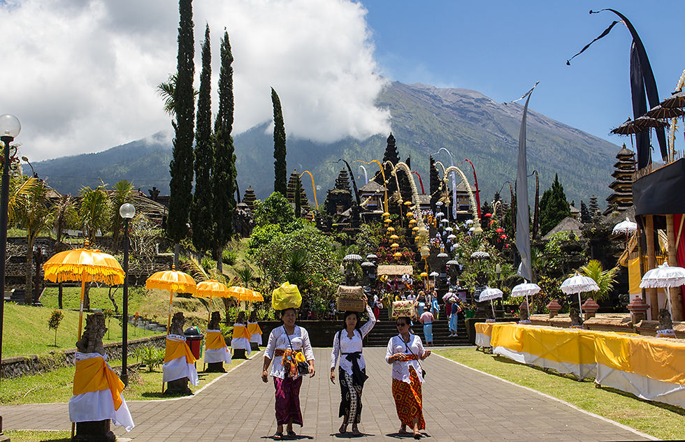 Women in ceremonial dress walk along a temple path lined with yellow umbrellas, with Mount Agung and clouds behind.
