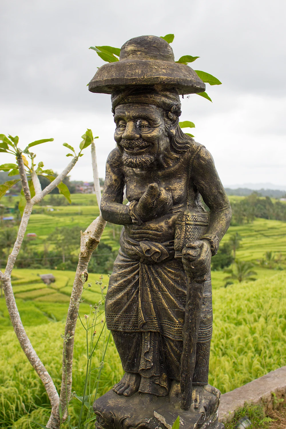 Ancient stone statue of a smiling man with a hat and staff in lush green rice paddy field; cloudy sky in the background.