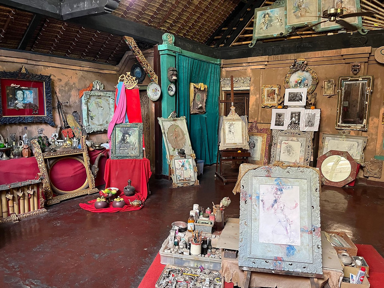 Art studio with framed sketches and paintings, vintage objects, and vibrant red and teal drapes. Rustic background and warm ambiance.