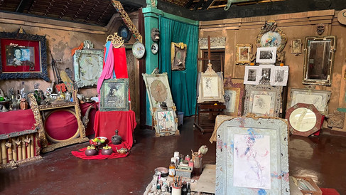 Art studio with framed sketches and paintings, vintage objects, and vibrant red and teal drapes. Rustic background and warm ambiance.