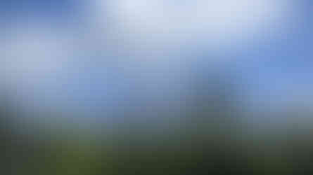 Volcano peak releasing smoke under a blue sky with fluffy clouds. Lush green trees in the foreground create a serene landscape.