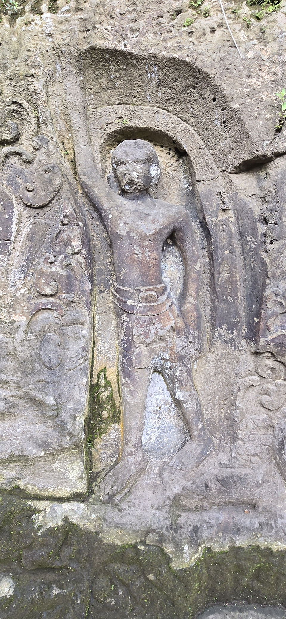 Weathered stone relief of a standing human figure carved into a rocky cliff, with moss and ornate patterns in an outdoor setting