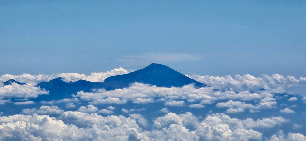Agung volcanic peak rising above a sea of white clouds under a clear sky, taken from a plane window