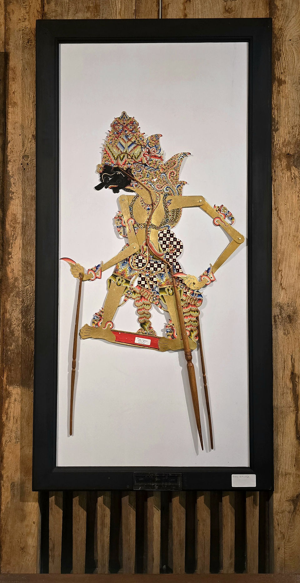 Intricately designed shadow puppet with colorful patterns on a white background, framed in black. Set against a wooden backdrop.