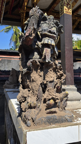 Weathered stone guardian statue under an ornate temple pavilion, dusty and eroded, with palm trees and bright daylight.