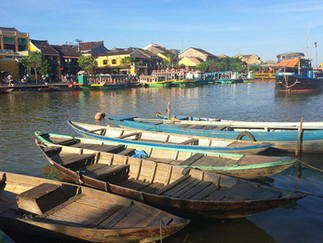 Wooden boats float on a sunny river with colourful buildings and larger boats docked in the background, creating a tranquil scene.