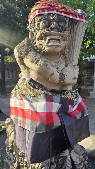 Stone guardian statue wrapped in red-check cloth and patterned sash, standing outdoors in sunlight with trees behind.