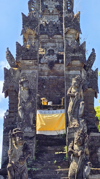Ornate stone temple gate with carved guardian statues and stairs, set against a bright blue sky; small offerings at the entrance.