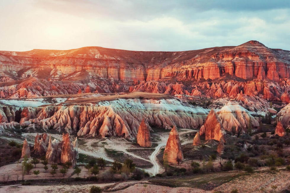 Rocky landscape at sunset in Cappadocia, Turkey. Eroded red and orange rock formations with a dusty path and scattered trees. Serene mood.