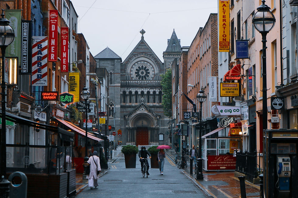 People walk and cycle on a rainy street lined with shops and neon signs, leading to a stone church in the distance. A vibrant urban scene.