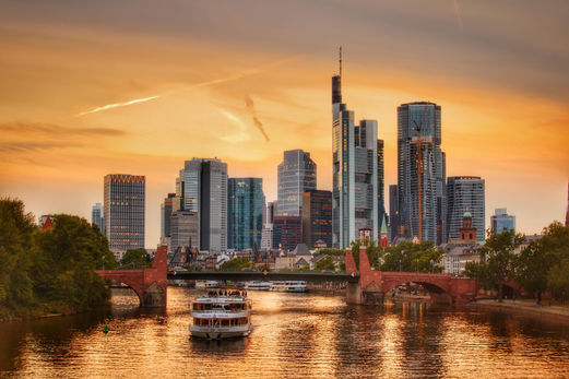 A scenic view of Frankfurt's skyline at sunset, with a river cruise boat gliding under a historic bridge, showcasing a captivating blend of modern skyscrapers and classic architecture along the riverbank.