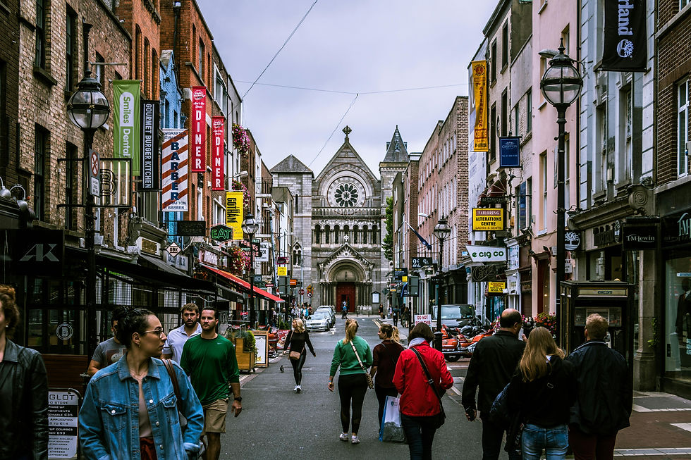 Busy street scene with people walking, surrounded by colorful shop signs and historic buildings. A large stone church is in the background.