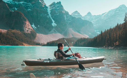 A kayaker paddles peacefully on the serene turquoise waters of a mountain lake, set against the backdrop of towering, snow-dusted peaks and lush evergreen forests.