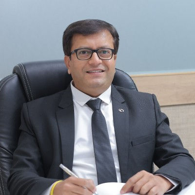 Smiling man in a dark suit and glasses sits at a desk in an office, holding a pen, with a calm professional mood.