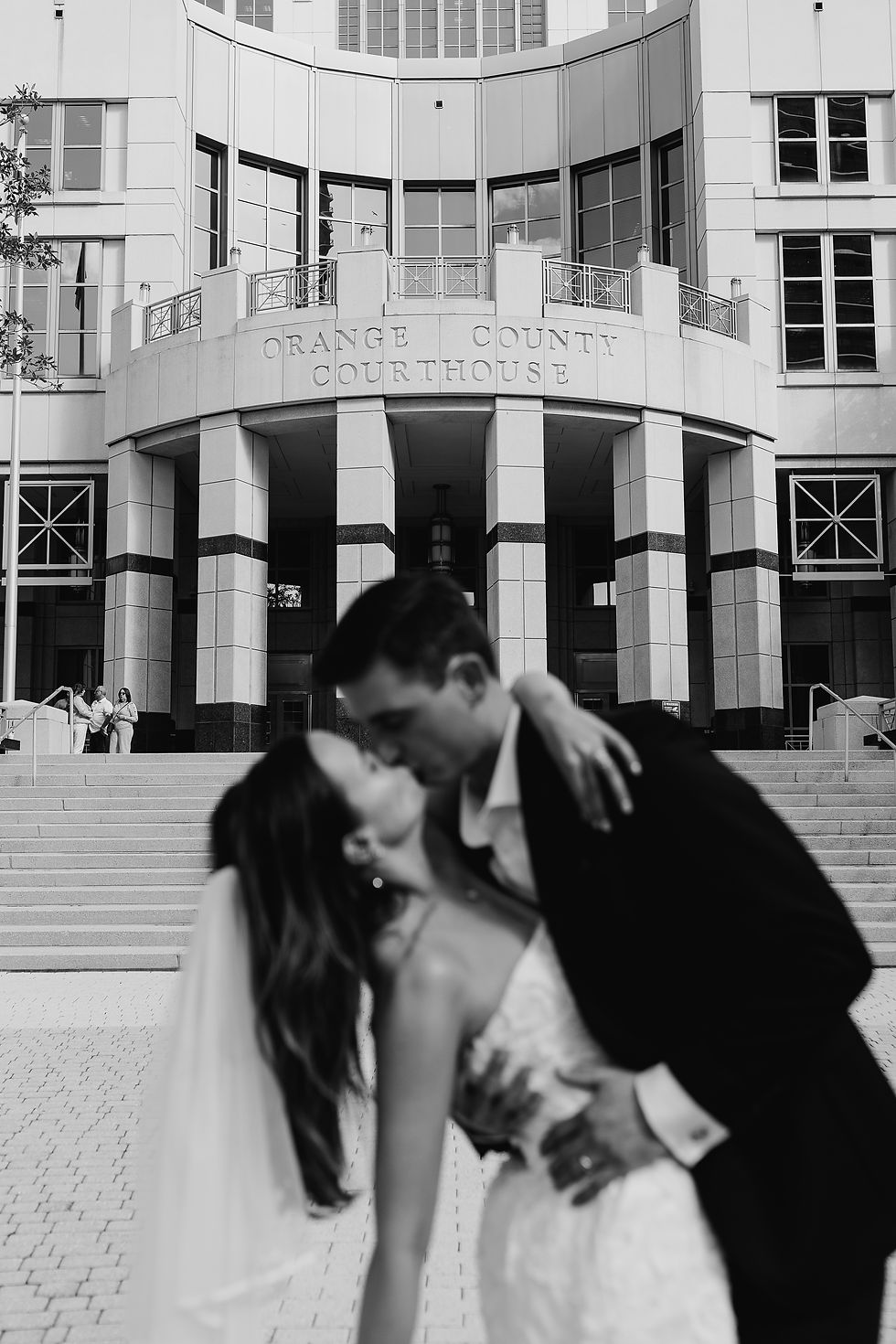 A couple kisses outside Orange County Courthouse. Bride in white dress and veil, groom in suit. Background shows courthouse entrance.