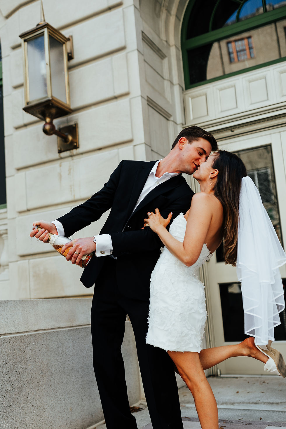 Couple in wedding attire kisses passionately. Groom in black suit pops champagne, bride in white dress and veil. Elegant building background.