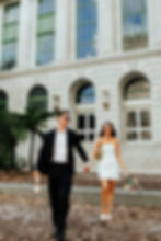 Couple in wedding attire holding hands, smiling, walking outside a grand building. Man holds champagne, woman carries bouquet. Sunny day.