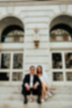 Bride and groom in sunglasses sit on steps, relaxed, with a bottle, bouquet nearby. Background: elegant building with large windows.