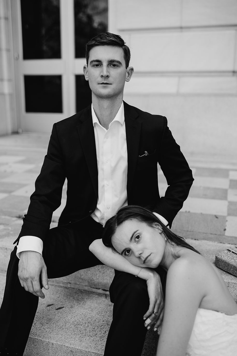 Man in suit sits on steps with a serious expression, while a woman in a white dress rests her head on his knee, looking contemplative. Black and white.