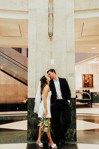 a man and woman in wedding attire lean against a marble column together and share a kiss.