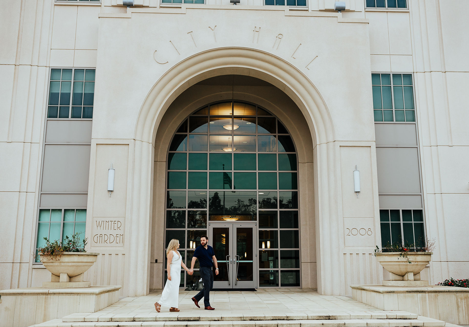 A man leads a woman across the steps in front of a large building with a glass archway. The text "Winter Garden City Hall" is visible.