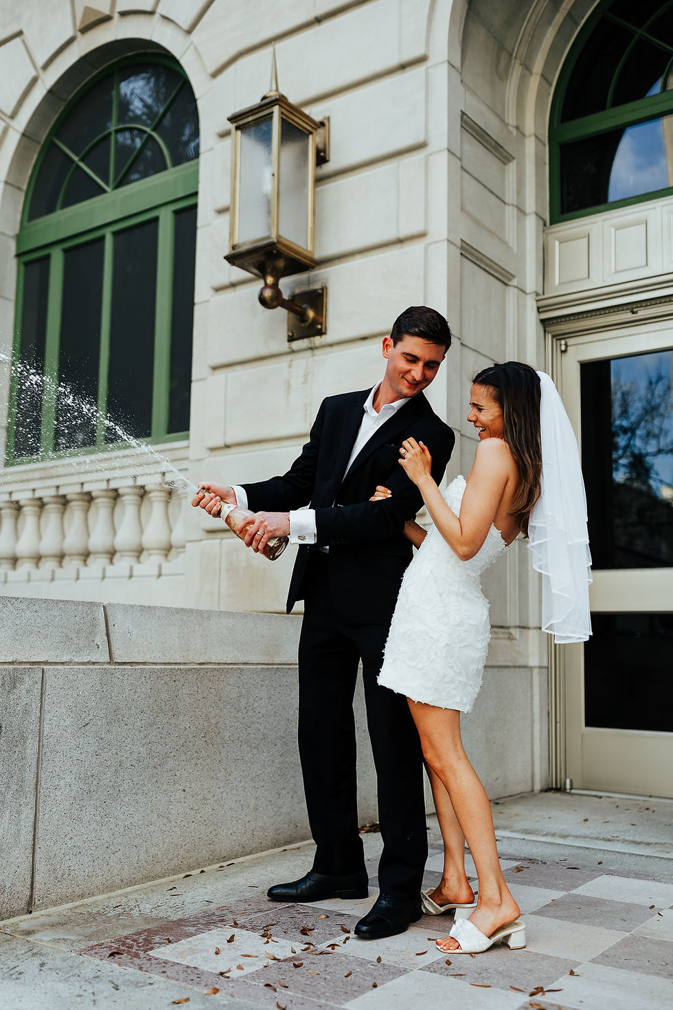 A smiling couple in wedding attire celebrate by popping champagne outside a stone building with arched windows and green trim.