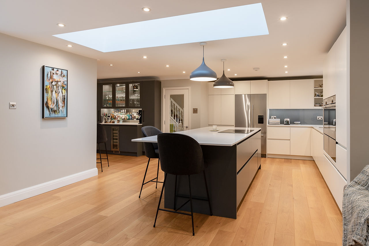 The handleless kitchen design features clean lines that not only make it aesthetically pleasing but also very easy to clean. The layout is simple yet ergonomically sound, with tall cabinets placed at both ends. A custom-made dresser-style cabinet with a sink is located on the left side, next to a built-in Fisher & Paykel fridge freezer. On the other end of the kitchen, there are ovens and a larder cabinet with a sink. Wall cabinets connect the two ends of the kitchen. The island has a Bora hob that is externally extracted, and ample drawer storage below for pots, pans, utensils, and other kitchen essentials.