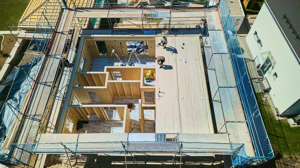 Drohnenaufnahme von dem Bau eines Holzhauses. Zwei Bauarbeiter stehen oben auf einem Holzflur.