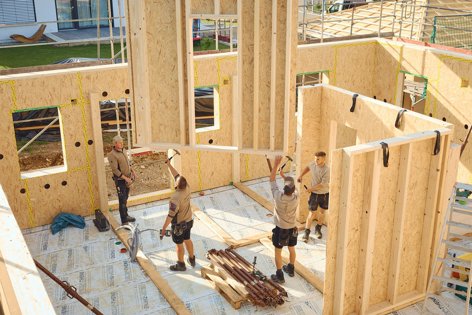 Vier Bauarbeitern befinden sich innerhalb eines Rohbau eines Holzhauses. Zwei Bauarbeiter arbeiten mit Hämmern an einer vom Kran hochgehobenen Holzwand. Währenddessen stehen zwei weitere Bauarbeiter (rechts und ganz links) an der Seite und beobachten die Situation.
