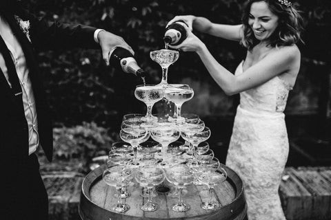 Black and white photo of a tower of champagne glasses on a wine barrel. A bride and groom are standing either side, smiling and pouring bottles of champagne into the top two tiers of the tower