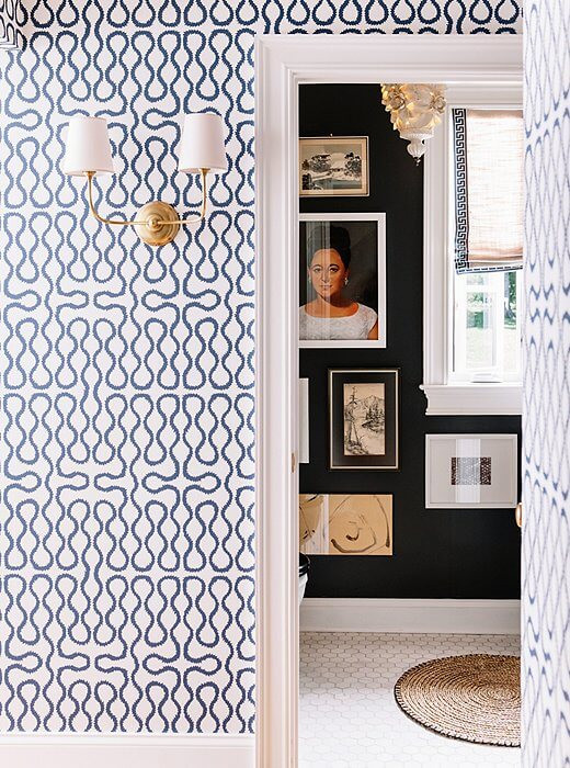 A wall is decorated with patterned blue and white wallpaper. A two-sided gold lamp is attached to the wall with simple white lamp shades. A doorway is visible on the right through which a black painted wall is visible with various paintings hanging on the wall. The floor is beige with a round jute rug