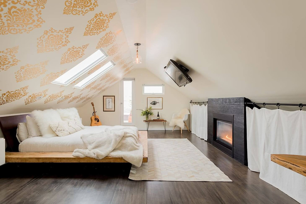 A loft style bedroom. The floor is a dark timber. The walls are beige; the ceiling is sloped to a point in the middle. The right ceiling slope matches the beige of the walls and has a TV mounted to it. The other ceiling/wall has gold damask wall paper with two large inset windows. A pendent light hangs from the middle of the roof. A bed sits in the middle of the room opposite the TV; it has white sheets with a wooden base and fluffy white cushions. Artwork hangs on the far wall with a clear-glassed door and a rectangular window. An acoustic guitar sits in a stand in the far corner. Opposite it there is a side table with a plant and a lush white arm chair. There is a fire place opposite the bed and a white rug rests at the foot of the bed