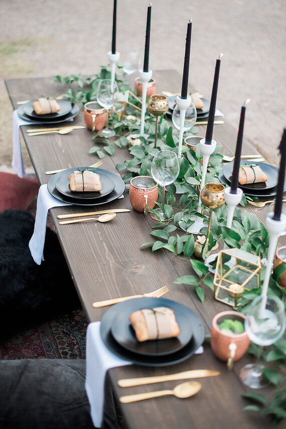 An overhead photo of a dark wooden table outside. The table has a eucalyptus garland runner with black tapered candles in white holders along the middle of the table. There are six place settings with black dinner plates and gold cutlery, and grey linen serviettes under the plates hanging off the edge of the table. On top of each place is a small wrapped gift in gold wrapping paper. Traditional wine glasses and pink and gold mugs rest at the top of each place setting