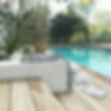 An outdoor pool and deck. In the foreground there is a staircase in light timber decking leading down to a decked area and pool on the right. The pool is rectangular and filled with turquoise water. There is a cream garden edge next to the stairs. A black and cream coat lays on top with a straw handbag. The garden has a fern and a few smaller cactus style plants and is filled with white pebbles. In the background there is a clear pool fence with lots of green trees and shrubs beyond.