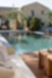 An outdoor pool. In the forground is a cream bench seat with beige cushions and a timber deck style table. The pool is surrounded by white tiles and timber decking and filled with clear-turquoise water. In the background is a sandstone house with native trees and a curtained cabana