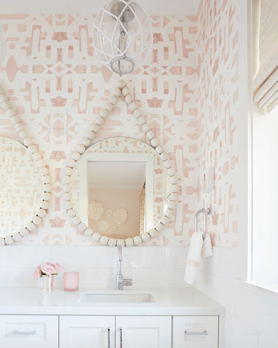 A white bathroom vanity sits against a wall with white tiles above and on the adjacent wall. On the wall above the vanity is a soft blush pink and white wall paper in an aztec style pattern. Two circular mirrors hand on the wall surrounded by cream wooden beads. a simple silver hand towel rail is attached to the wall on the right with a white handtowel hanging from it. There is an open window to the right and blush pink accessories on the vanity. A small orb shaped light hangs from the ceiling with white macrame decor on the outside of the glass