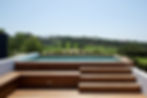A photo of an outdoor pool. In the foreground there is timber decking and a bench seat/storage space. Timber stairs lead up to an above ground pool. It is surrounded by the same timber decking with white retaining walls either side. The pool is filled with turquoise water and overlooks a majestic view of green hills, tan gravel paths and lots of trees.