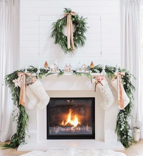 A wood fire burns in a white fireplace. The chimney and walls are painted white and a soft white fur rug rests in front of the fire on a light toned wooden fllor. A pine garland hangs over the mantelpiece, hanging down the sides. Candles sit on top of the mantel and four white knitted stockings hang from it with rose gold coloured ribbons. A matching pine wreath with a rose gold ribbon hangs on the chimney above the mantel