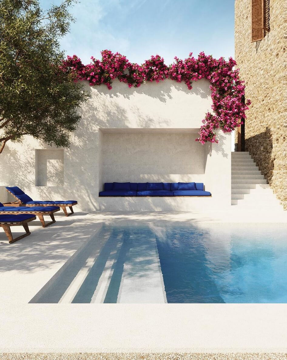An outdoor pool and entertaining area. The area is paved in cream with large steps leading into a pool on the right. The pool is filled with clear-blue water. On the left of the pool are two brown deck chairs with blue cushions. A tree overhangs. Behind the pool there is an inlaid alcove with a blue bench seat. Pink vined flowers drape over the top of the alcove. A set of stairs leads behind the pool to the right of the alove. The side of a tan bricked building is seen next to it.