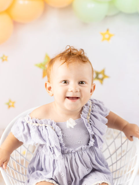 A one year old little girl sits in a chair with a rainbow balloon arch backdrop behind her during a studio photo shoot.